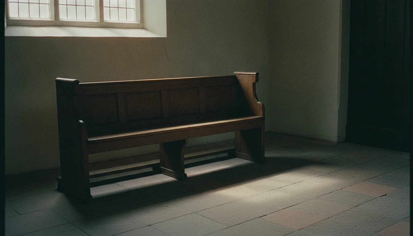 An empty wooden pew in soft light from a tall plain glass window, the rest of the room in shadow.